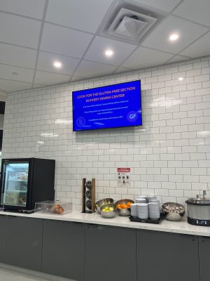 Digital sign promoting gluten-free section at university dining center, above a food counter with bowls, plates, and utensils.