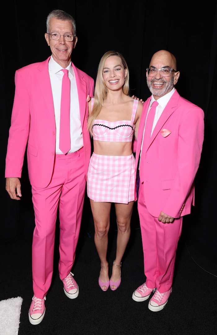 Three people smiling at a formal event, dressed in pink. The person on the left is wearing a pink suit and glasses, the middle person is in a striped pink and white dress, and the person on the right is in a pink suit and white shirt.