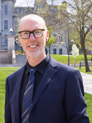 A person in a dark blue suit with a smile, standing outdoors with green grass and a historic building in the background.