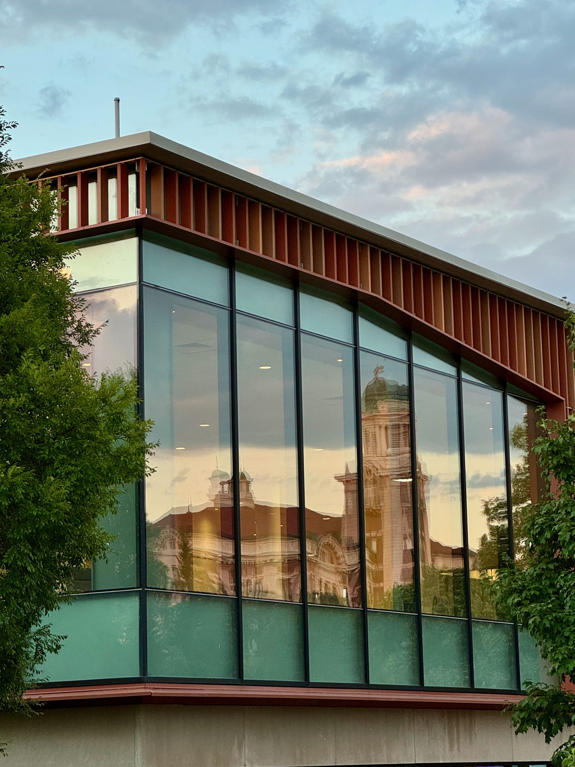 Exterior view of a modern building with large glass windows reflecting an image of a traditional building with a prominent clock tower, set against a dusk sky.