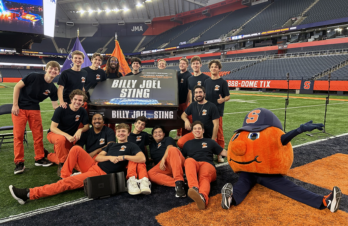 A group of people wearing matching black shirts and orange pants are posing around a “Billy Joel/Sting” sign on a football field. They are smiling and accompanied by Otto the Orange mascot. A large stadium is in the background.