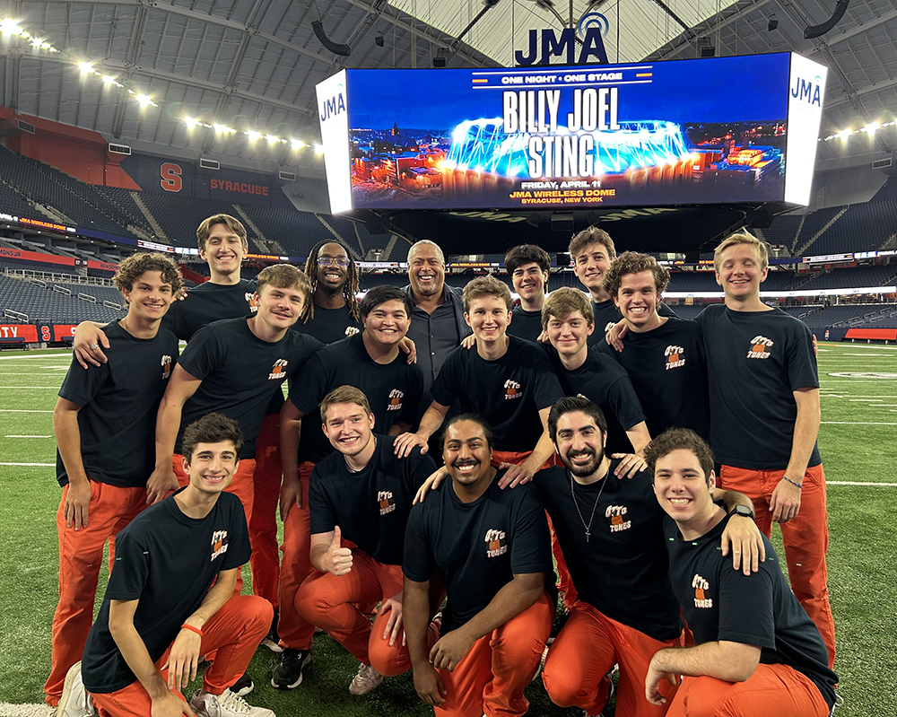 A group of 17 people, dressed in matching black T-shirts and orange pants, smiles for a photo on the field inside the JMA Wireless Dome. A large screen behind them displays a concert announcement for Billy Joel and Sting. The venue and other details are visible in the background.