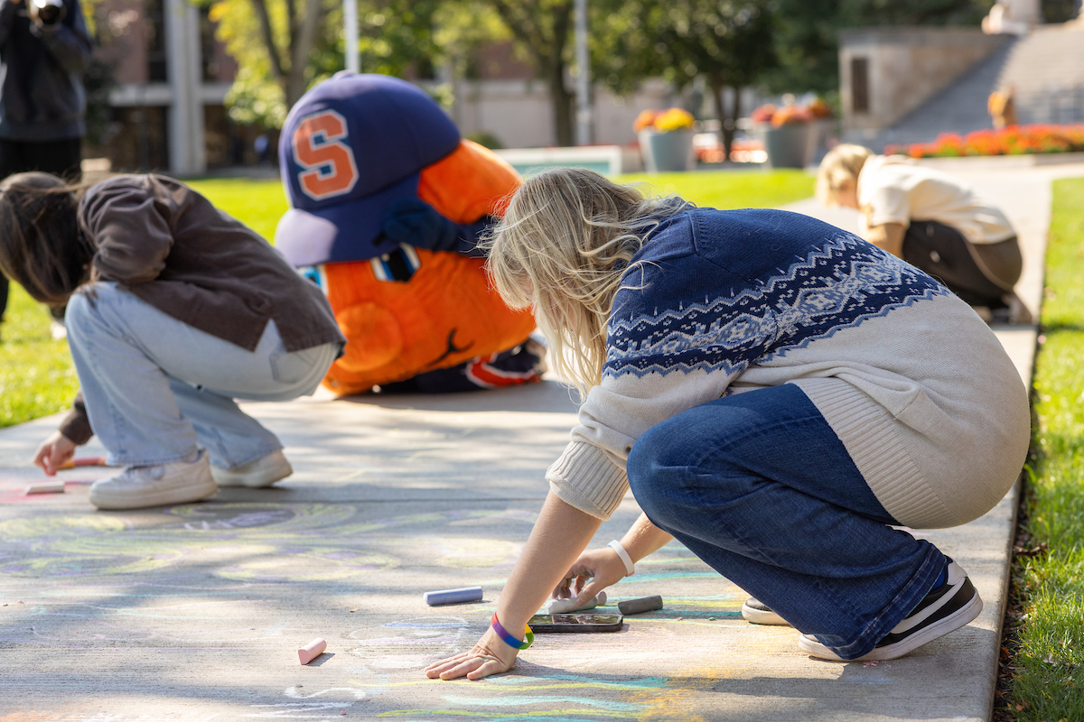 Students, including the Otto the Orange, drawing with chalk on the Quad's sidewalk on a sunny day.