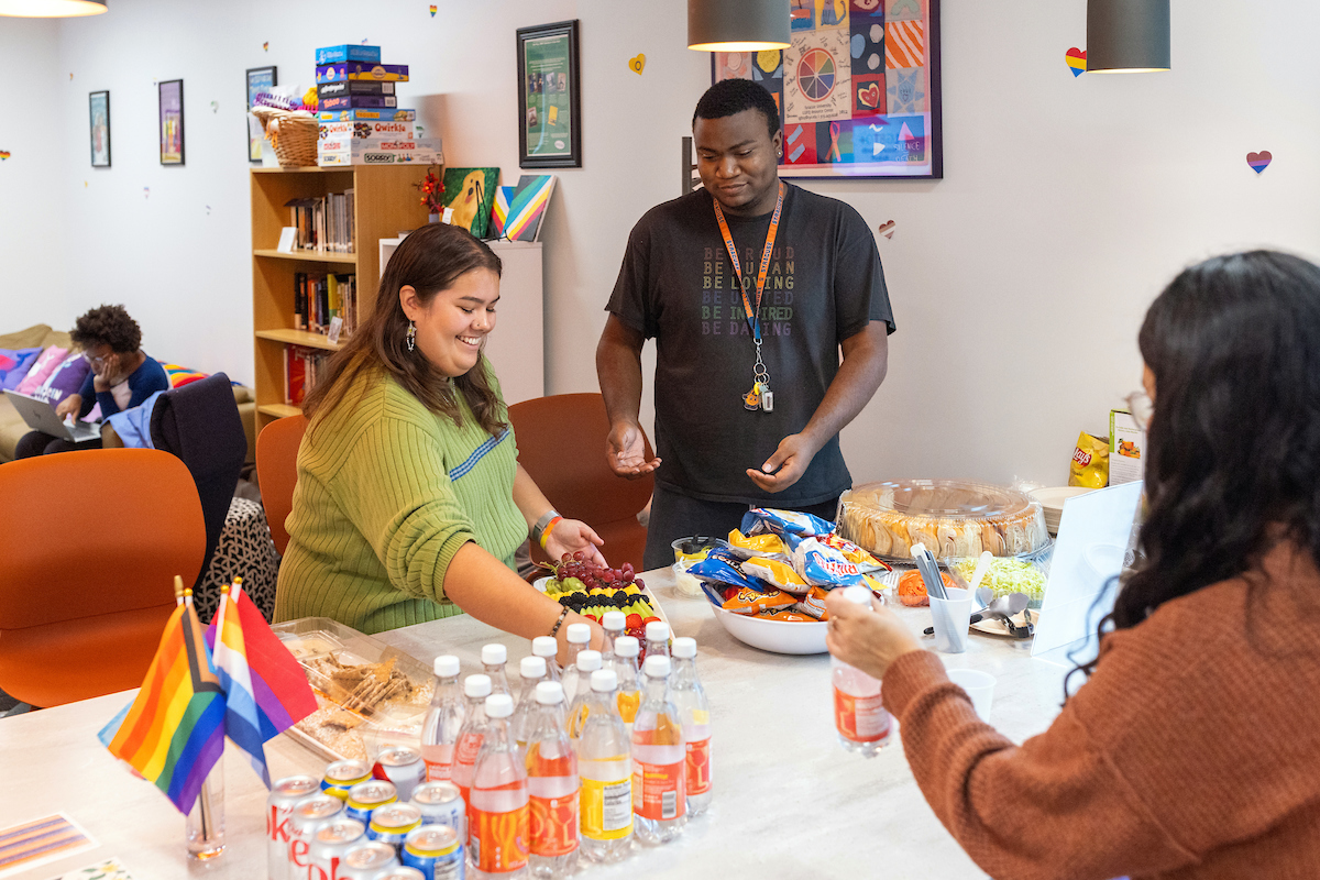 Three individuals enjoying a casual office gathering with snacks and beverages on a table, featuring a visible pride flag.