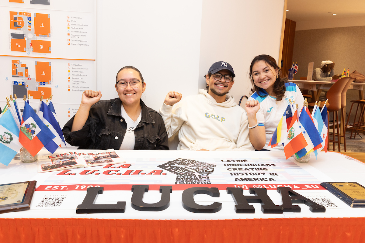 Three students seated at a table smiling and holding their fists up at a table promoting La LUCHA with multiple countries' flags