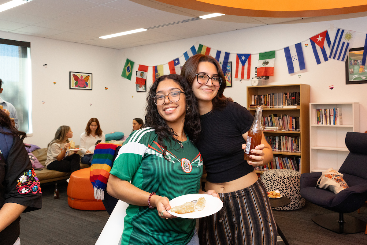 Two students hold up food and drinks and smile together at an event celebrating Latine Heritage Month. Colorful flags from Latin nations appear in the background