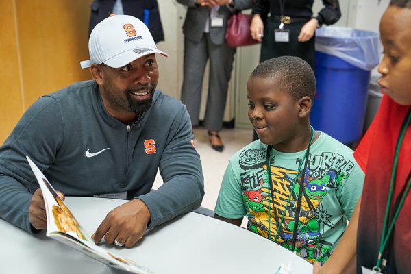 A seated man in a Syracuse cap and zip-up jacket is showing a book to a young boy in a Teenage Mutant Ninja Turtles T-shirt. The boy is smiling, and they are seated at a table. Another child stands nearby, attentively watching.