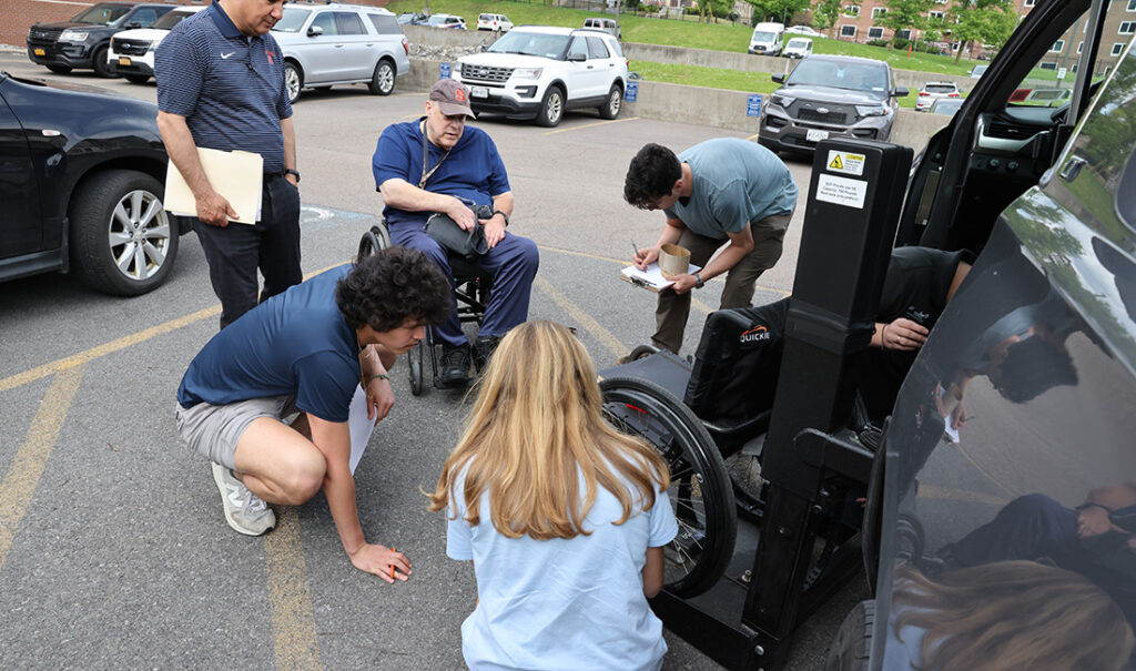 Five individuals, including one in a wheelchair, gathered around an empty wheelchair that is attached to a vehicle. 