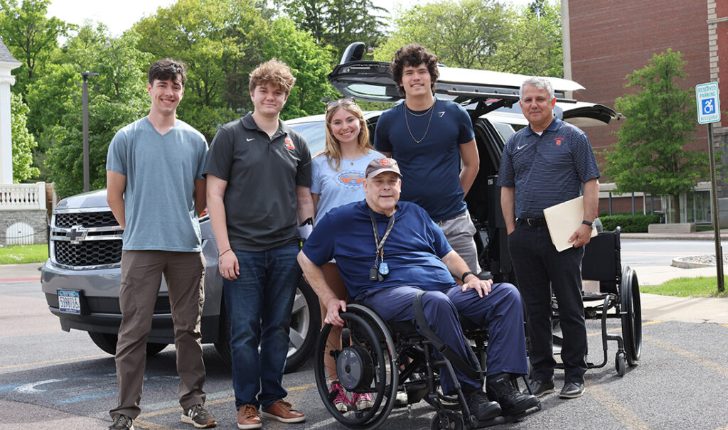 Five people standing in front of a car gathered around a person sitting in a wheelchair.