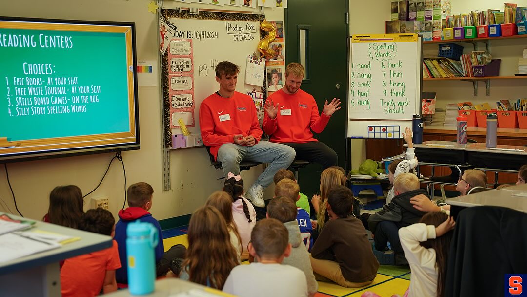 Two individuals in orange shirts reading to a group of children seated on the floor in a colorful classroom filled with educational posters and reading materials.