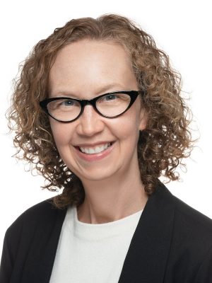 A person with chin-length blonde curly hair and black glasses wearing a black jacket and white blouse