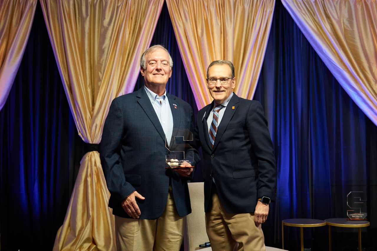 Two individuals posing with an award at a formal event, standing in front of draped golden curtains. The person on the left is wearing a suit with a pin and the person on the right is also in a suit, wearing glasses.