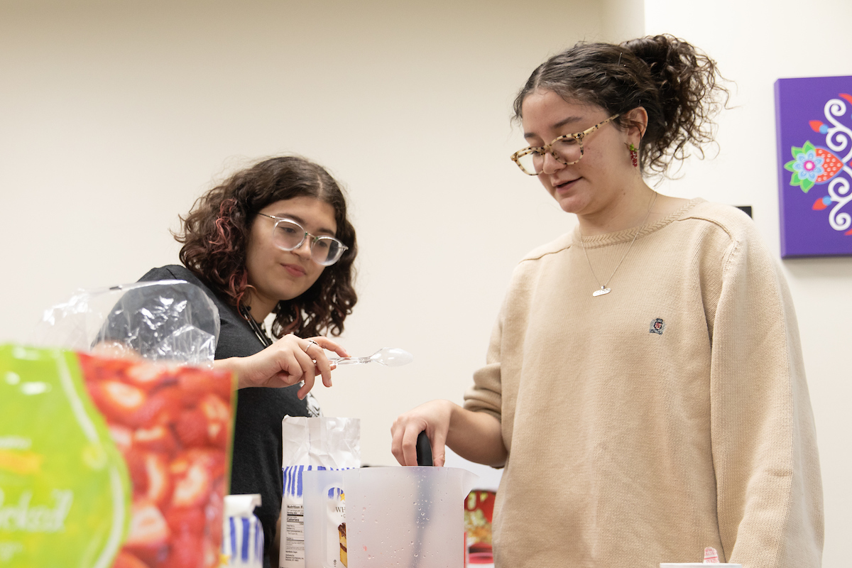 Two individuals interacting while making juice, with one person holding a spoon and the other is measuring out sugar. Various food items and kitchenware are visible on the table.