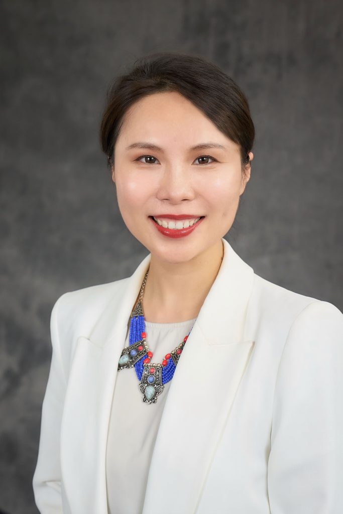 Portrait of a smiling professional wearing a white blazer and a colorful necklace, set against a gray background.