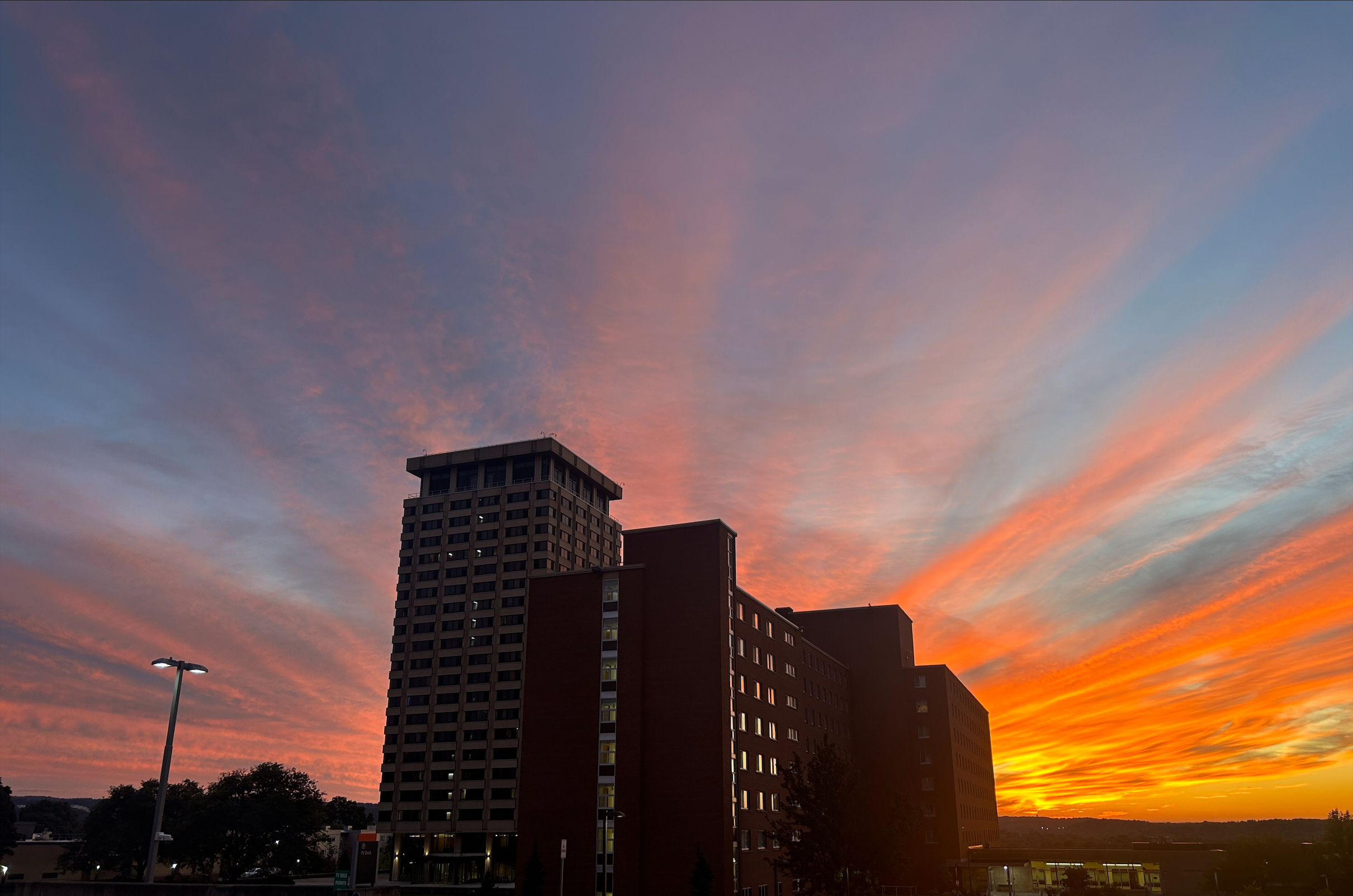 A vibrant sunset with streaks of orange and pink clouds over a city skyline featuring several high-rise buildings.