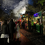 A bustling evening scene at an outdoor festival with people browsing colorful food stalls and a firework exploding in the sky.