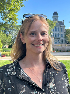 A smiling person with the Hall of Languages at Syracuse University in the background on a sunny day.