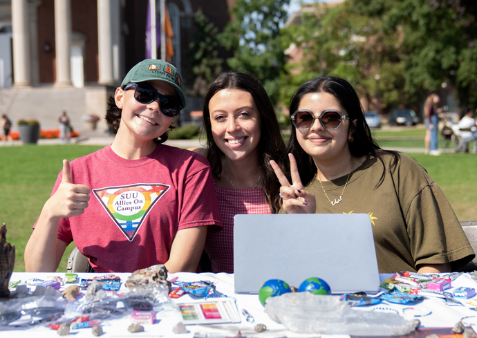 Three students smile while seated at a table on the Quad as part of the student involvement fair.