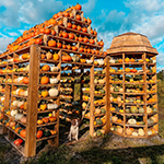 A dog standing in front of an elaborately structured pumpkin tower featuring multiple varieties of pumpkins and gourds, set against a backdrop of blue sky and clouds.