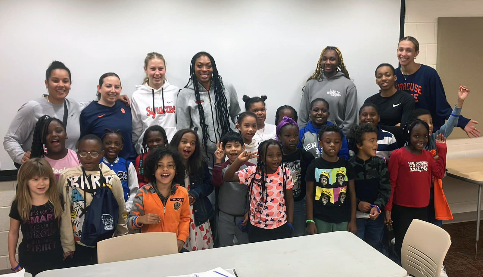 A group of people comprising elementary school children and college-aged student athletes pose together in front of a whiteboard