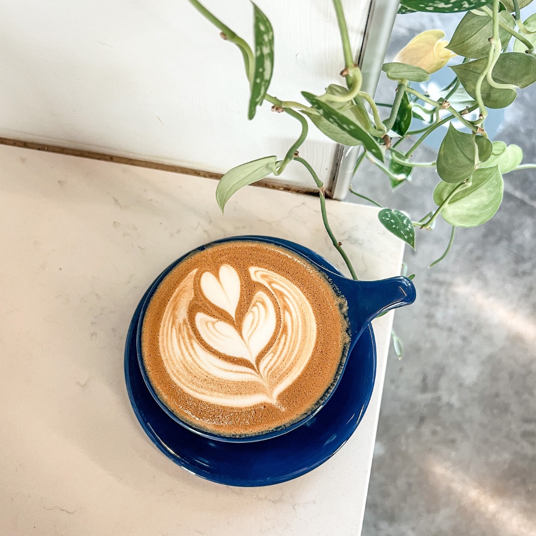 A cup of cappuccino with a latte art design of a heart on top, placed on a white surface next to a hanging green plant. The cup is blue and uniquely shaped with a broad base and curving handle.