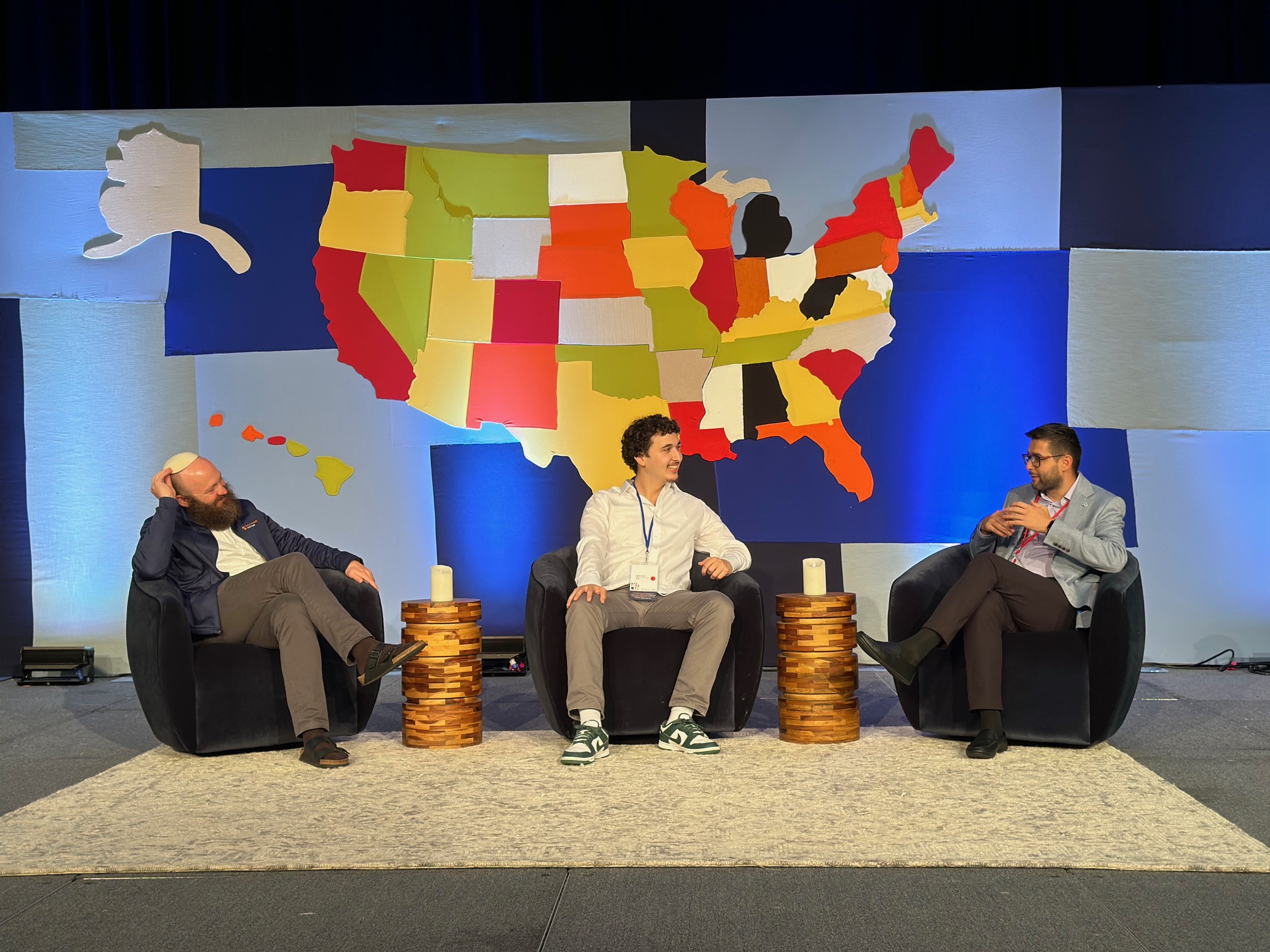 Three individuals are seated on stage in front of a large, colorful map of the United States, engaging in a discussion. The stage features unique stool-like structures and each participant wears a name tag and casual business attire.