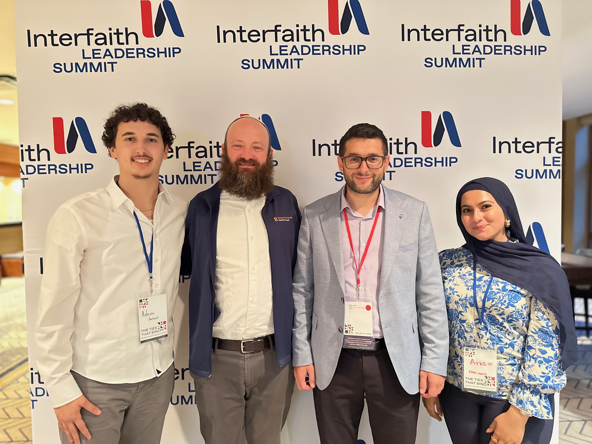 Four individuals smiling at an Interfaith Leadership Summit, standing in front of a banner with the event's name. They are wearing conference badges, and one person is wearing a hijab.