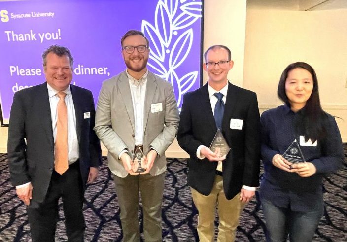 group of four people, three holding awards, in front of a screen that includes the Syracuse University logo and the words "Thank you!"