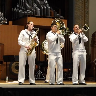 Five naval musicians perform with brass and woodwind instruments in a concert hall with a pipe organ backdrop