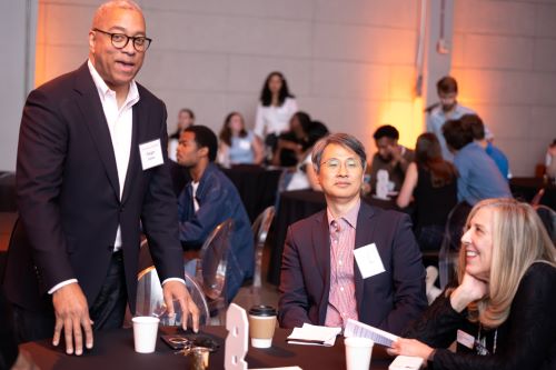 man standing and a man and woman seated at a table at a reception