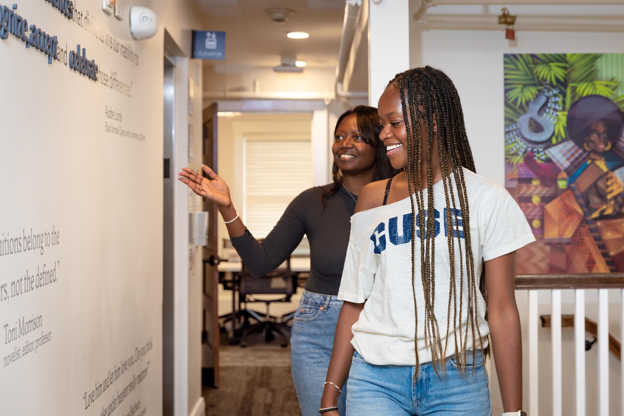 Two individuals smiling and walking through a hallway with motivational words on the wall. One person is pointing at the wall text as they engage in a conversation.