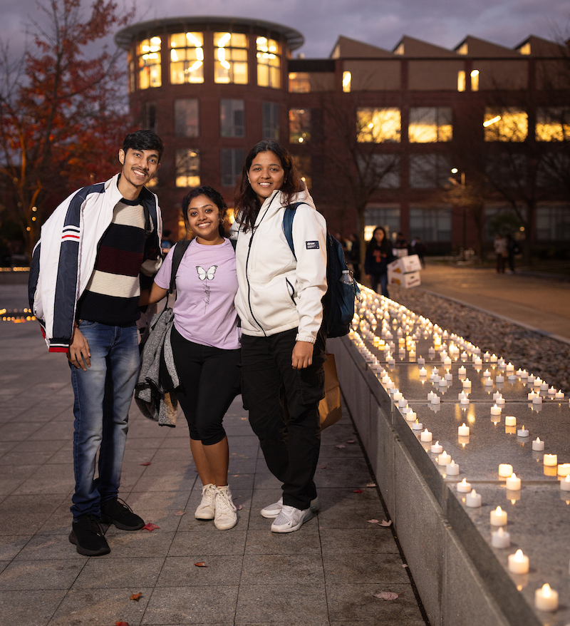 Three students smiling in front of a campus building in the evening, lined with lit candles.