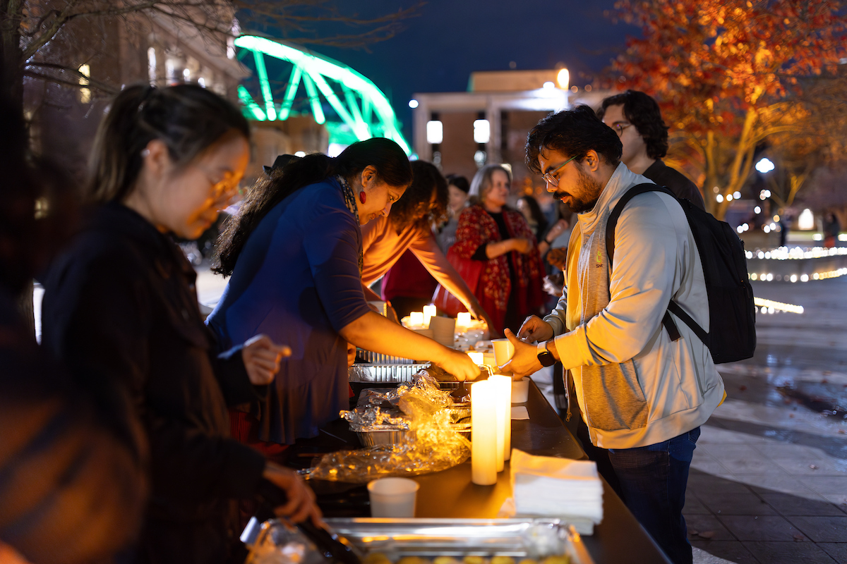 People serving and receiving food at an outdoor evening event, with decorative lights and the JMA Wireless Dome in the background. 