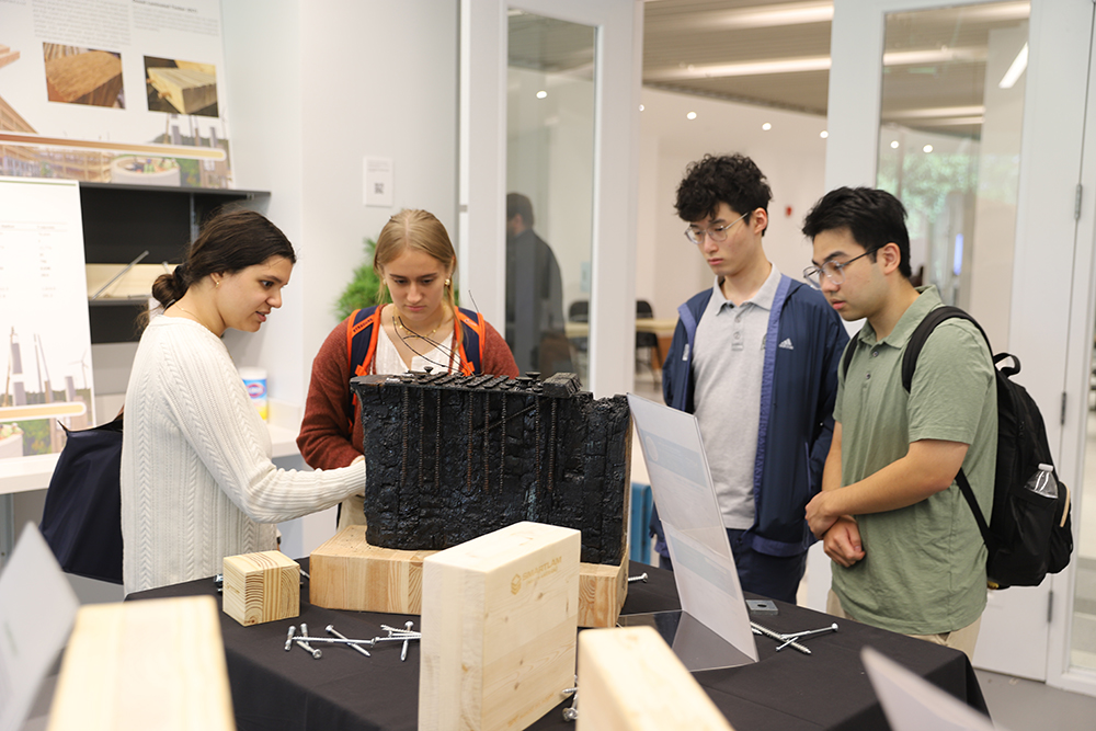 A group of four students examines a dark architectural model placed on a wooden base, displayed on a table in a classroom. Posters with architectural designs are visible on the walls in the background.