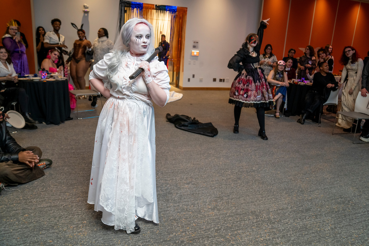 Person in detailed white costume with face painted white and dark eye makeup stands at the foreground in a room with an audience, while another individual in a black and red outfit performs in the background.