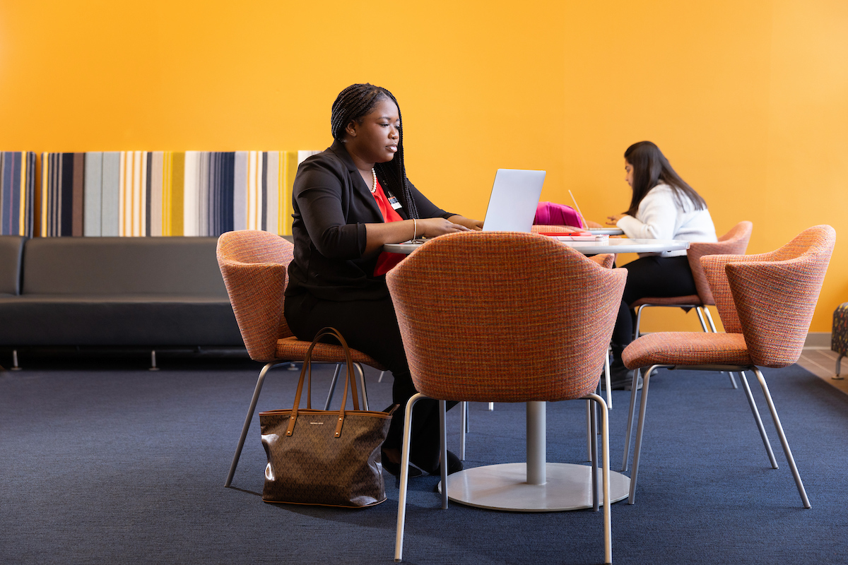 Person working on a laptop at a table in a vibrant workspace with yellow walls and modern furniture. Another individual is seated in the background.