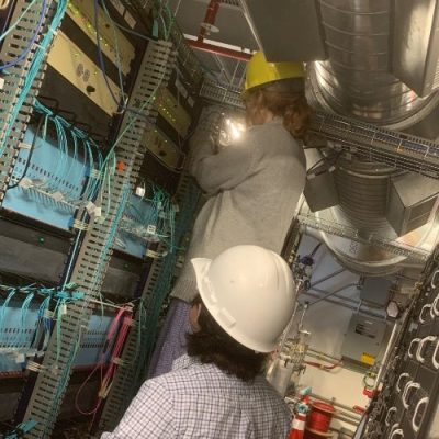 Two individuals in hard hats are inspecting and working on network equipment in a server room.