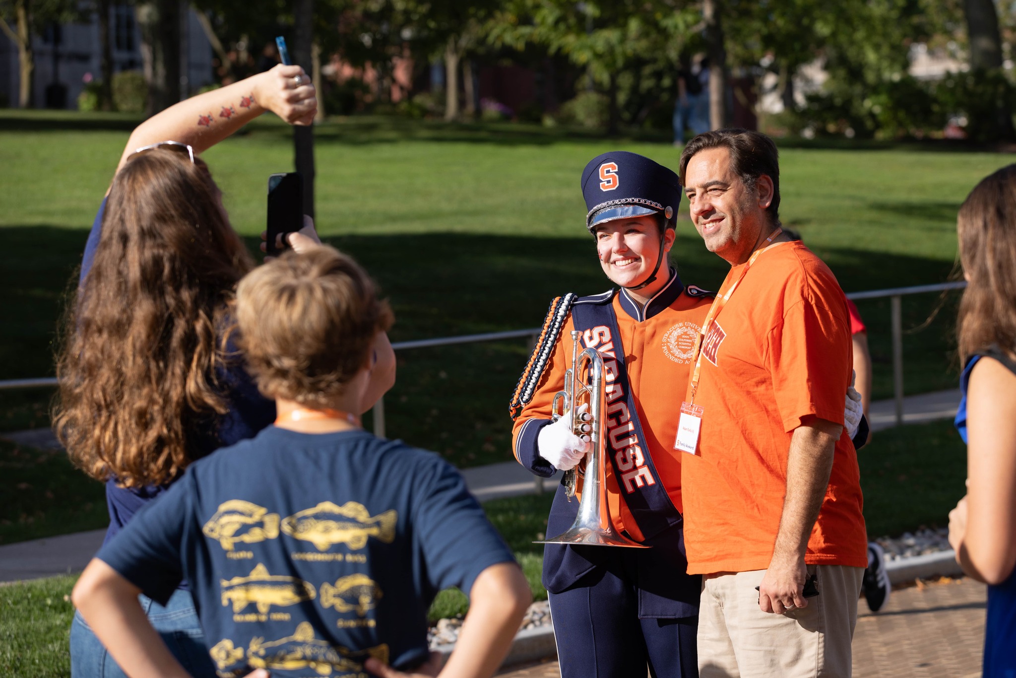 Person in Syracuse University marching band uniform takes a selfie with another individual wearing a Syracuse shirt, while others observe, outdoors on a sunny day.