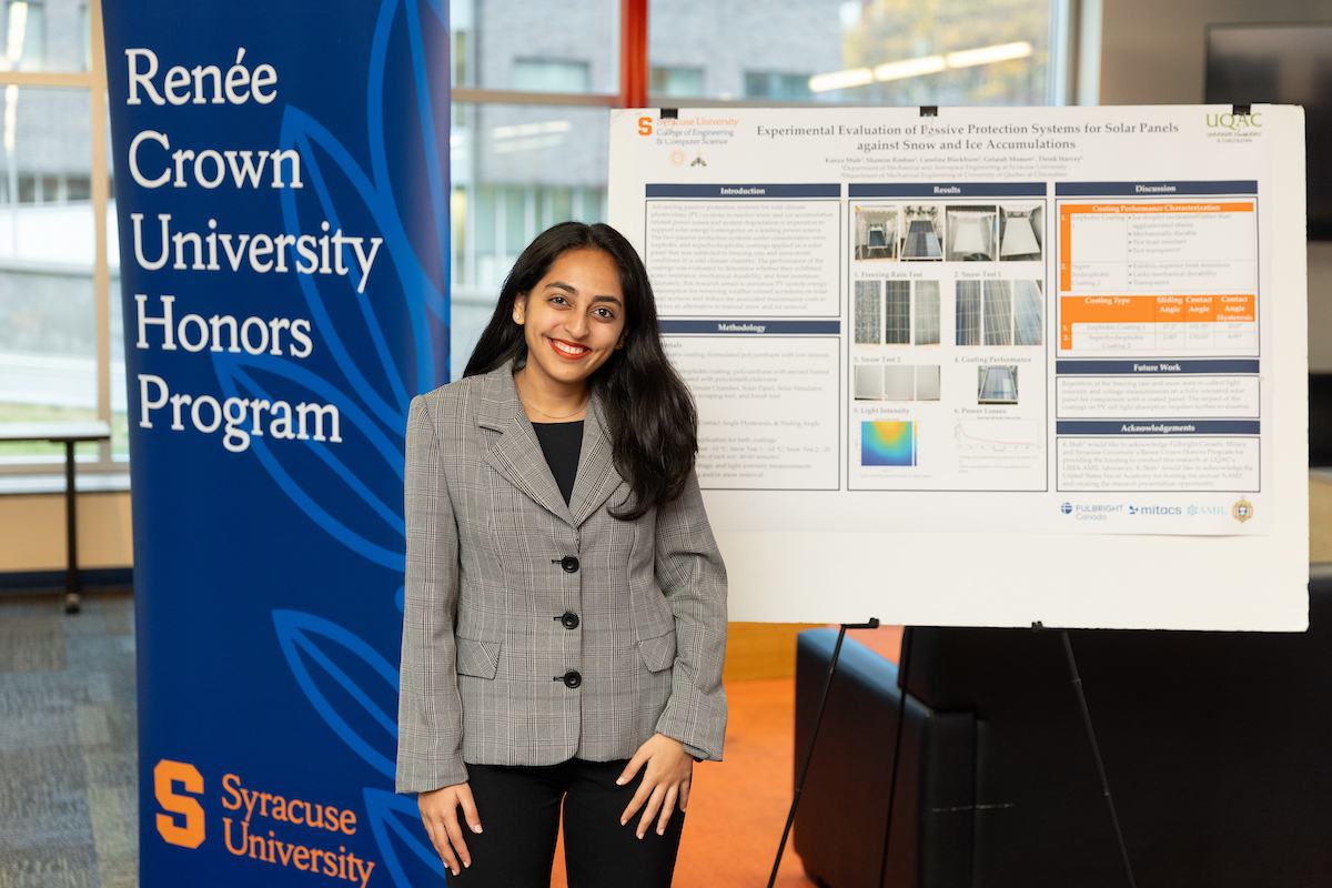 A person standing between two banners, one reading "Renée Crown University Honors Program" and the other displaying a research poster titled "Experimental Evaluation of Finite Precision Systems for Safe Roads" at Syracuse University. The individual is smiling and dressed in business attire.