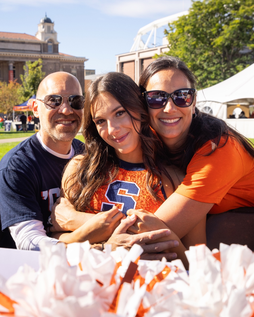 Three people smiling and embracing at an outdoor event with a university building and tents in the background. They wear clothing with the logo of Syracuse University.