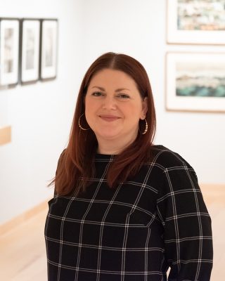 A person with long reddish-brown hair, wearing a black plaid top and hoop earrings, stands in an art gallery with framed artwork on the walls. They are smiling and facing the camera.