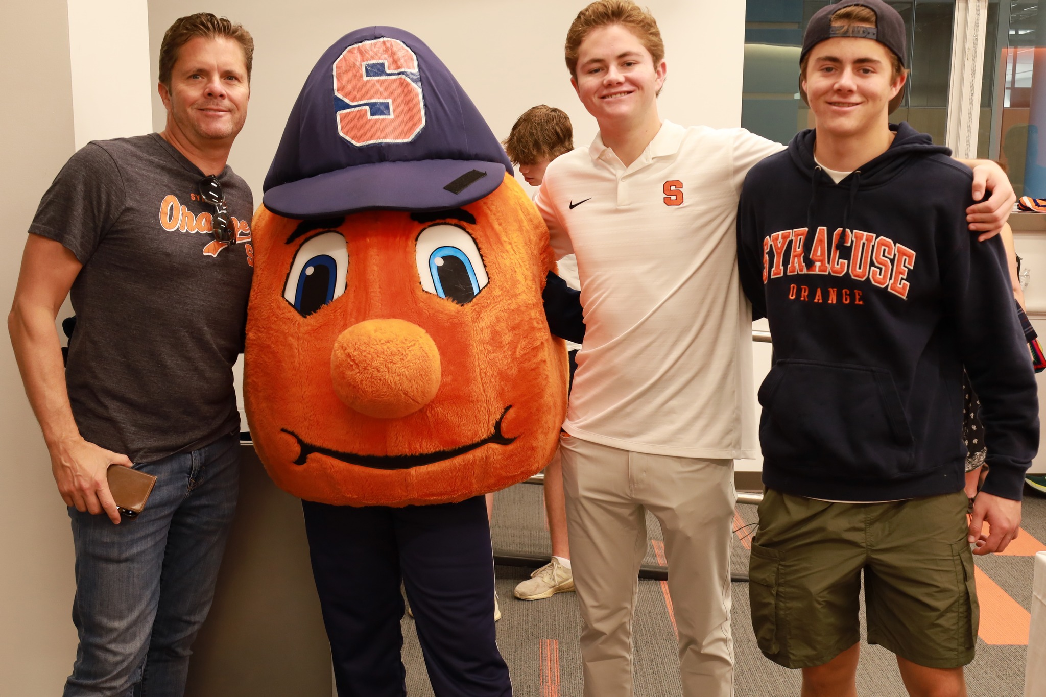 Three people posing with the Syracuse Orange mascot at an event, two wearing Syracuse University apparel.