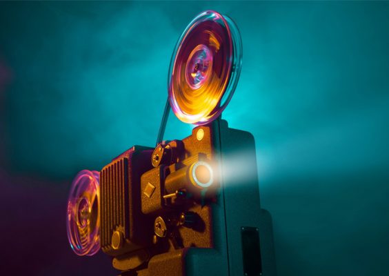 A vintage film projector with spinning reels casts a beam of light, set against a vibrant background of blue and purple smoke.