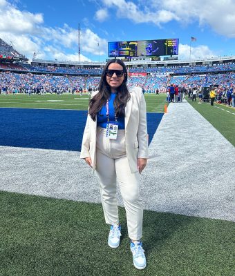 A woman in a white suit and blue top stands on a football field near the end zone. She wears sunglasses and sneakers. The stadium is filled with fans, and a big screen is visible in the background under a partly cloudy sky.