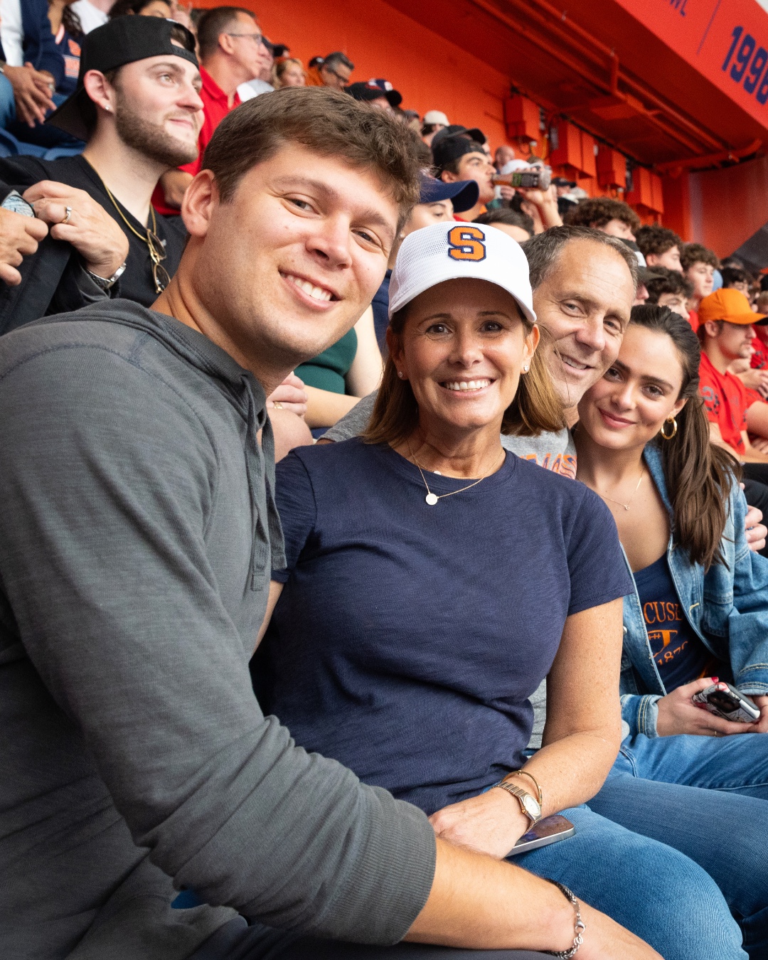 Four fans sitting in stadium seats at a Syracuse University sports event, smiling for the camera.