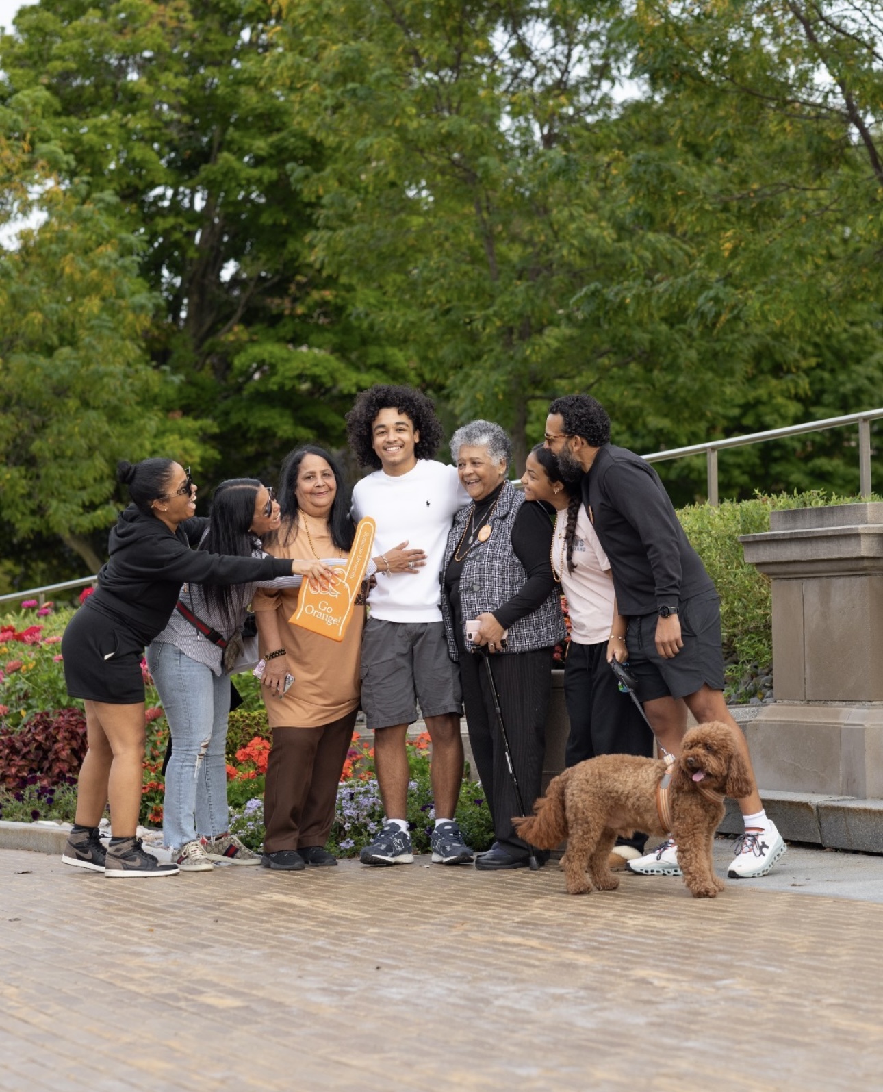 A group of six people smiling and embracing in an outdoor setting with lush greenery and flowers around them.