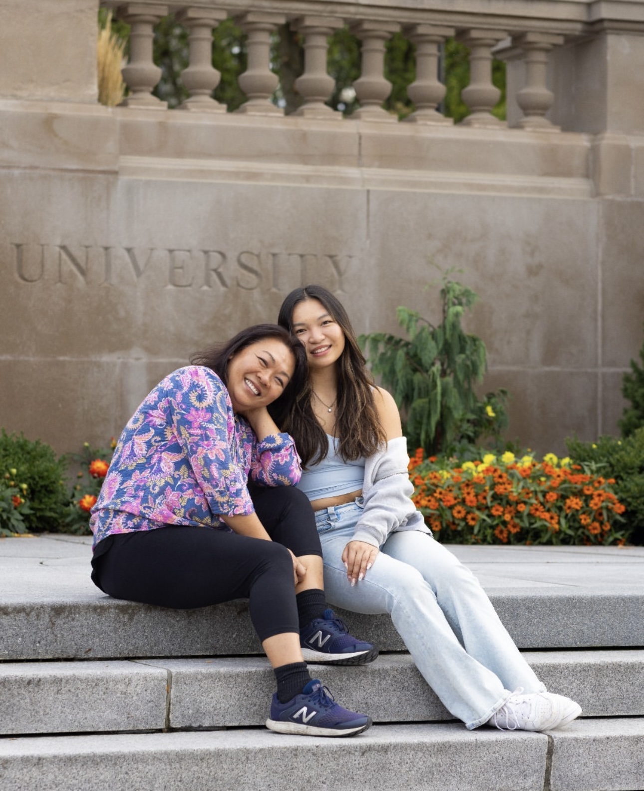 Two individuals are sitting closely together on the University's steps, smiling at the camera. One is wearing a floral shirt and black pants, and the other is dressed in a gray top with light blue jeans. 