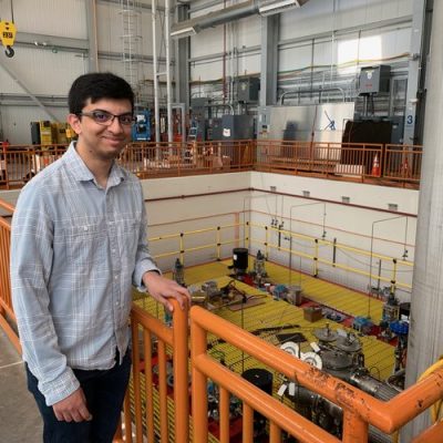 Person smiling at the camera, standing by a railing with an industrial setting featuring large machinery and equipment in the background.