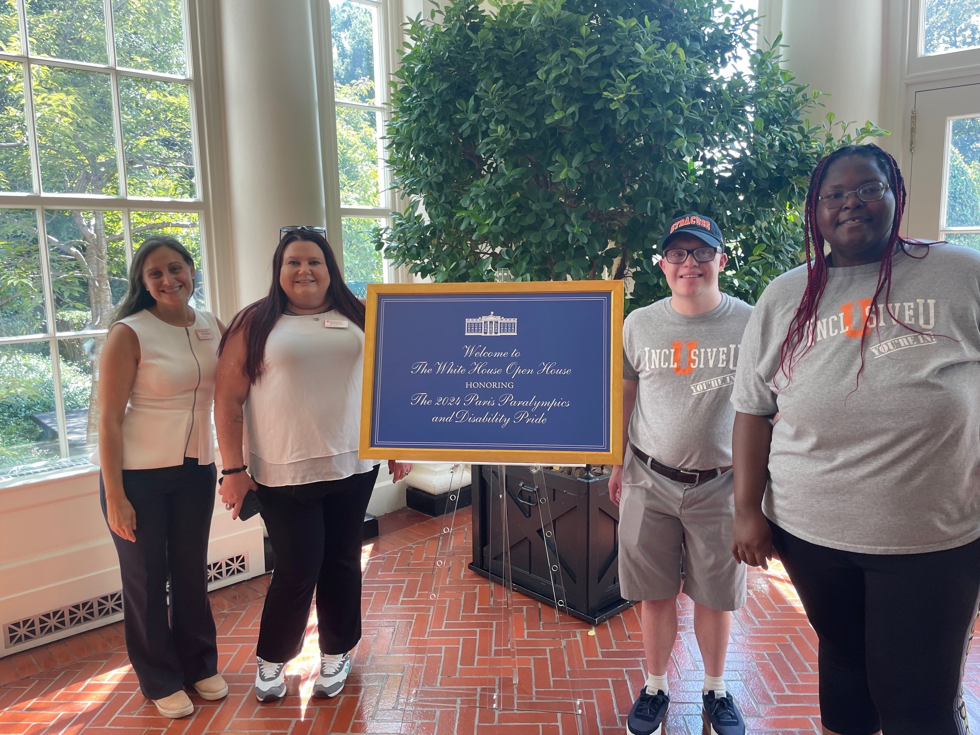 Two staff members and two students pose before a sign reading Welcome to the White House open house honoring the 2024 Paris Paralympics and Disability Pride.