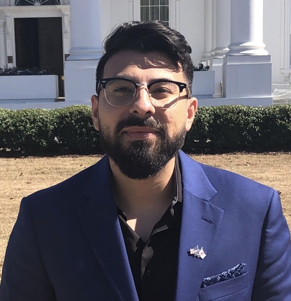 An individual wearing glasses and a blue blazer stands in front of the Atlanta White House on a sunny day.